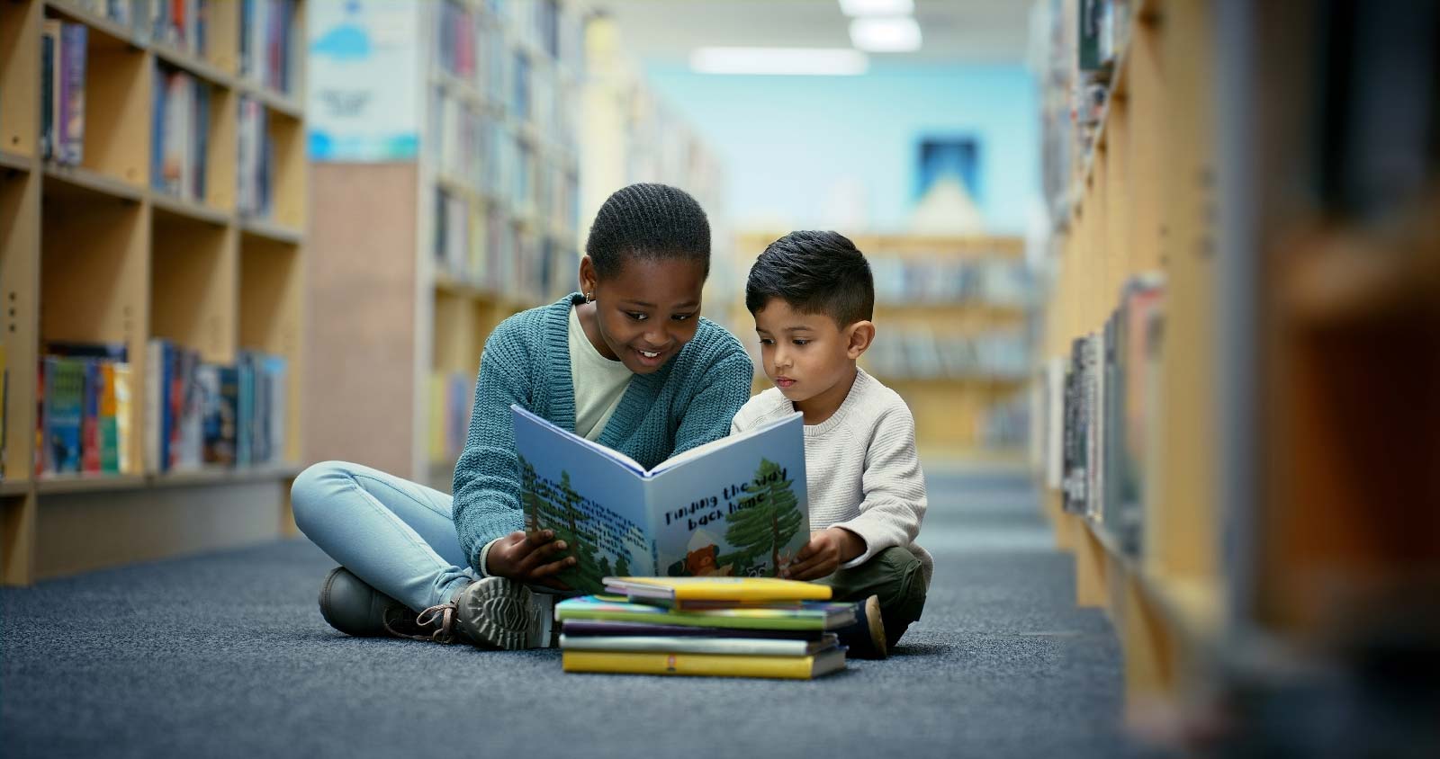 A young girl and boy sitting on the floor of a library reading a book together.